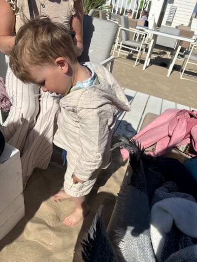 The image captures a relaxed outdoor scene, likely a beachside terrace or cafe, with sand covering the ground. The setting appears to be a sunny day, indicated by the bright light and distinct shadows cast across the sand and furniture. Based on the provided context, this location is Middelkerke, Belgium.
In the foreground, a young child with light brown hair is partially visible, appearing to rest or gaze downwards. The child wears a light beige or off-white, textured long-sleeved outfit, possibly a linen jacket, over a light blue and green patterned undershirt, and has a pacifier in their mouth. Their bare feet are visible, resting on the sand. The child is close to an adult, whose arm and upper torso are visible, wearing a shimmering, light-colored top with a necklace.
Various items are scattered around the seating area. To the right, there's a vibrant pink suede-like bag or jacket, alongside several blankets or throws in shades of grey, white, and black, some with striped or patterned textures and fringed edges. The furniture in the background consists of modern, white metal chairs and tables, suggesting an outdoor dining or lounge area. The presence of sand directly underfoot, combined with the furniture, points to an upscale beach club or terrace rather than an open beach. No visible text is present in the image.