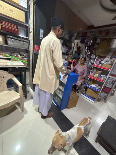 The image depicts the interior of a retail establishment in Ambernath, India, likely a pet supply store. In the foreground, a light brown and white dog stands on a white tiled floor bisected by a black strip. A male individual, positioned with his back mostly to the camera, stands near a blue, narrow counter or display stand. He wears a light-colored long shirt (kurta), white trousers, dark sandals, and a black cap. He is looking towards the blue stand, which displays "Pay PLEASE" and "REMOVE YOUR FOOTWEAR OUTSIDE" text. A brown cardboard box rests at the base of this stand.

Behind the blue stand, a female individual is seated, facing towards the left. She wears a patterned purple dress and holds a white object in her left hand. To the left of the frame, a light-colored plastic chair and a dark table are visible, featuring papers and a green broom handle. Behind these items, shelving units contain various products, including boxes labeled "VET-PRO" and assorted containers. To the right of the scene, multiple white metal display racks hold packaged products, several marked "SIZE HEALTH NUTRITION," along with a variety of pet accessories hanging on a wall grid. A QR code is visible on a dark panel in the background. The ceiling shows a fan.