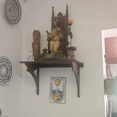 The image shows a wall with several decorative items, including ceramic plates, a wooden carving, a statue of Jesus, and a sundial tile. One ceramic plate, with a blue and white floral pattern, is mounted on the wall to the left. Below it, another similar plate is partially visible.
The central focus is a wooden shelf mounted on the wall. On the shelf, from left to right, are a small white ceramic bell with blue designs, a wooden carving of a human head with braided hair, a statue of Jesus seated on a throne holding a green orb and a white object in his hands, and a glass vase with dried flowers. A single artificial rose with green leaves is placed behind the statue.
Below the shelf, a rectangular ceramic tile depicting a sundial with a sun face, a village scene, and Roman numerals is affixed to the wall. The wall itself is textured and painted white. To the right of the shelf, a portion of a doorway or window opening is visible, with a wooden frame. A rope is tied to a bracket near the opening. A red fire extinguisher is partially visible in the background, near the doorway.
This scene is consistent with decorations found in homes or small establishments in Lloret de Mar, Spain. The decorative plates, religious imagery, and sundial are common elements in Spanish homes and tourist areas, reflecting a blend of cultural and religious traditions.