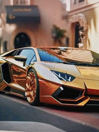 The image shows a close-up of a gold Lamborghini Aventador parked on a paved surface, possibly a street or driveway. The car has a sleek, aggressive design with sharp angles and a low profile. Its gold paint is highly reflective, mirroring the surroundings. The front of the car features distinctive headlights and air intakes. The rear wheel, with its intricate gold rim, is prominently visible. In the background, a building with an arched doorway and architectural details suggests a European setting, but there is no indication of the specific city of Sluck, Belarus. The lighting suggests it is daytime. There are no people or any other discernible objects or activities in the frame.