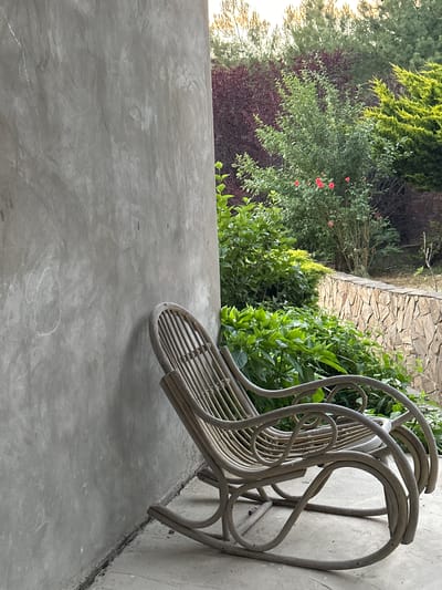 This image showcases an outdoor scene, likely a patio or balcony in Myskhako, Russia, with a focus on a rattan chair leaning against a concrete wall. The chair, made of light-colored rattan, has a distinctive looped design on its backrest and a slatted seat. It appears to be unoccupied, suggesting a moment of stillness or rest.
Behind the chair, a lush green garden thrives. Various types of foliage are visible, including dense bushes with bright green leaves and taller trees with needle-like branches. In the mid-ground, small red flowers, possibly roses, add pops of color to the greenery. The garden appears well-maintained and vibrant.
A stone pathway or wall runs diagonally across the upper portion of the image, adding a textural contrast to the softer organic shapes of the plants. The ground in the foreground is a smooth, light grey concrete surface, which shows some subtle marbling and a faint white outline of an unknown shape.
The lighting suggests it might be late afternoon or early morning, with a soft, diffused light illuminating the scene. The overall mood is tranquil and serene, capturing a quiet moment in a natural setting. No people or visible text are present in the image.