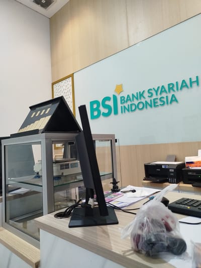 This media file captures an indoor scene at a Bank Syariah Indonesia (BSI) branch in Lhoksukon, Indonesia. The setting appears to be a customer service counter or teller area.
In the foreground, a wooden counter is visible, on which a black computer monitor stands prominently. Cables extend from the monitor into a hole in the counter. To the right of the monitor, a keyboard and a transparent plastic bag filled with what appear to be red and dark-colored items are placed on the counter. Papers and a key are also scattered on the counter.
Behind the counter, a glass display case is partially visible, containing some items and a scale. On top of the display case sits a black, pyramid-shaped object with gold-colored panels.
The background features a wall with wooden paneling and a light blue section with the BSI logo. The text "BSI BANK SYARIAH INDONESIA" is clearly visible in teal lettering on the light blue wall. To the right, two printers are placed on a shelf.
The lighting suggests it is daytime. There are no people visible in the image, creating a sense of quiet or being between customers. The overall impression is a functional banking environment.