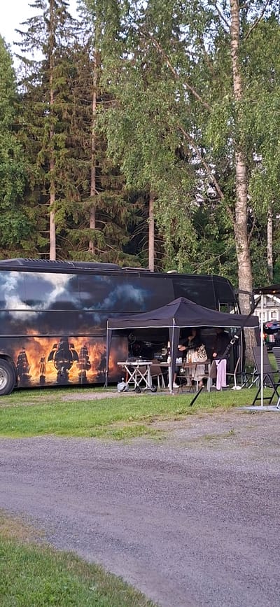 The image shows a black bus with a graphic of sailing ships on fire against a cloudy sky. In front of the bus is a black canopy tent set up over a table with chairs. Several people are standing and interacting under the tent, some holding cups or drinks. A portable grill and a gas cylinder are visible on the table. The scene is outdoors, with a gravel path and green grass in the foreground, and a dense forest of pine and birch trees in the background. There are also some white structures and chairs visible to the right of the canopy. The location appears to be a campsite or an outdoor event venue in Salpakangas, Finland.