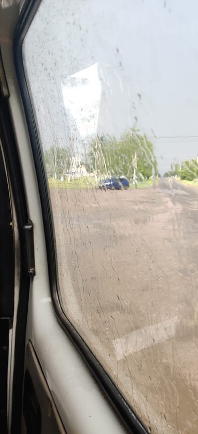 The image shows a view through a rain-streaked window of a vehicle, likely in Shelestove, Ukraine. Outside, a dirt road is visible, with a blue car parked on the side. Trees and some buildings can be seen in the background. The sky is overcast, indicating rainy weather. The interior of the vehicle, specifically the door and window frame, are visible on the left side of the image.