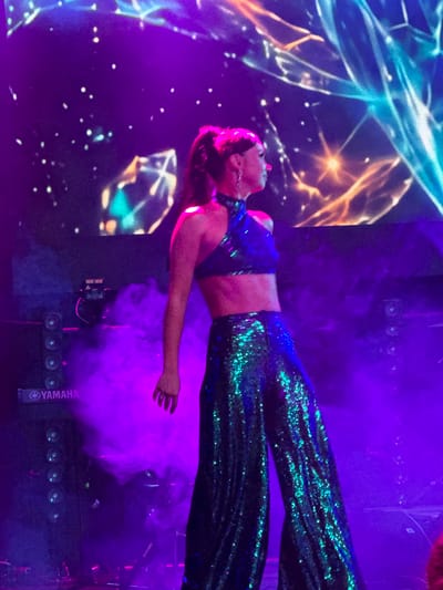 A full-shot, eye-level, medium-low angle medium-shot captures a performer on stage in Tsilkani, Georgia. The performer, a woman, is facing to the right of the frame, looking off into the distance with her head tilted slightly. She has on a sparkly blue halter top and matching wide-leg pants. Her hair is pulled back in a ponytail. A large screen behind her displays a vibrant, abstract image with purple, blue, and orange hues. The stage is illuminated by purple lighting and filled with smoke. To the left of the performer, a Yamaha logo is visible on a piece of equipment. There are multiple speakers stacked vertically next to the Yamaha equipment. The overall scene suggests a live music performance or concert.