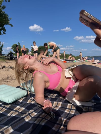 In Ust'ye, Russia, on a sunny day at the beach, a young woman lies on her back on a checkered blanket. She is wearing a pink bikini and a white skirt tied around her waist. Her head is tilted back, and her mouth is wide open in a laugh. Her eyes are closed, and her hair is falling around her face. Next to her is a light blue inflatable pillow. In the background, other people are scattered across the sandy beach and near the water, enjoying the warm weather. The sky is a clear blue with a few fluffy white clouds, and trees line the edge of the beach.