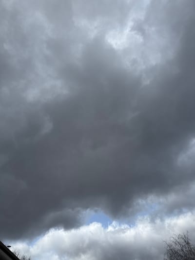 The scene depicts a sky heavily overcast with multiple layers of dark grey stratus and nimbostratus clouds, indicative of an impending or ongoing weather event. A narrow, irregular opening in the lower cloud layer, positioned towards the bottom-center, reveals a small segment of lighter grey clouds and a sliver of blue sky. In the bottom-left quadrant, a dark, angular structure consistent with a roofline or eave is partially visible. To the bottom-right, the bare, leafless branches of a deciduous tree extend into the frame against the lighter clouds. No individuals, explicit actions, or interactions are present. The environmental conditions suggest a day with low visibility and potential precipitation in Oberhausbergen, France.