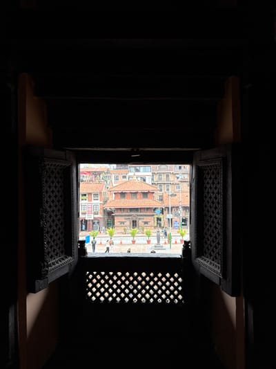 View from a dark interior through a window onto the architecture and street scene of Lalitpur, Nepal. The window has ornate lattice grills at the top, bottom, and to the right. Outside the window, buildings with brick roofs, whitewashed walls, and traditional architecture line a street scene, where people are walking. Potted plants and orange traffic cones are visible. The angle of the photograph suggests the view is from above street level. The room from which the photo is taken is dark, with visible structural details on the left and right.