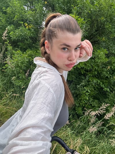 This is a selfie of a young woman lying down outdoors. She is wearing a white oversized shirt and grey leggings. Her right hand is touching her forehead, and her gaze is directed towards the camera. She has light brown hair styled in a ponytail and is wearing makeup, including lipstick.

The background consists of lush green trees and tall grass, suggesting a natural, possibly rural setting. The lighting indicates it is daytime, and the overall atmosphere appears relaxed and casual. The image is taken from a high-angle perspective, emphasizing the woman's face and upper body against the natural environment. There is a hint of a black object with a curved handle visible on the right side of the frame, possibly part of a stroller or bicycle. No visible text is present in the image. The implied location is Шевлягино, Russia.