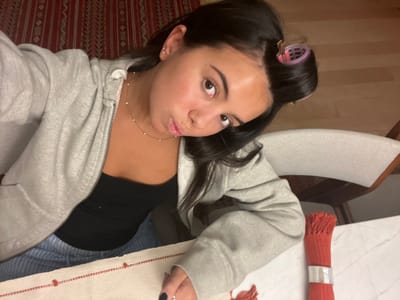 An overhead selfie captures a young woman seated at a table in an indoor residential setting in Lordelo do Ouro e Massarelos, Portugal. The woman, with dark hair, has a pink hair roller visible in a section of her hair on the right side of her head. She is wearing a light grey zippered hooded sweatshirt over a black strapless top and light blue and white striped bottoms. A delicate gold-toned necklace with small white beads is around her neck, and she has stud earrings in her left ear. Her lips are pursed into a "duck face" expression as she looks upwards towards the camera. Her left hand, with black-painted fingernails, rests on a white tablecloth adorned with a red striped pattern featuring small red heart-like motifs. On the white marble-patterned tabletop to her right, a rolled red textile item with fringed ends, secured by a silver ring, is present. Behind her, a dark red patterned textile, possibly a rug or wall hanging, is visible. To her right, a cream-colored upholstered chair with a dark wooden frame is partially visible, next to a light wood floor.