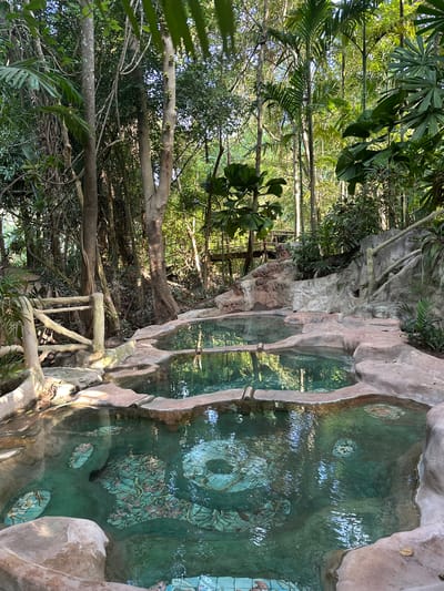 The image depicts a series of tiered, natural hot spring pools nestled within a lush, tropical forest environment. The pools are filled with clear, emerald-green water, and the bottom of the pools features intricate mosaic tiles depicting floral and natural scenes. The pools are constructed from natural rock formations and concrete, designed to blend seamlessly with the surrounding landscape.
The forest is dense with a variety of trees, including palm trees and other tropical vegetation, creating a shaded and serene atmosphere. Sunlight filters through the canopy, casting dappled light on the water and surrounding rocks. A rustic wooden railing and some stone pathways are visible, suggesting a developed tourist area. In the background, a wooden bridge or walkway can be seen elevated among the trees.
The location is identified as Khlong Thom Nuea, Thailand, a region known for its natural hot springs. There are no people visible in the image. The scene is tranquil and undisturbed, highlighting the natural beauty of the hot springs and their jungle setting.