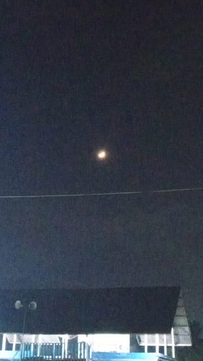A nighttime photograph captured from below, looking upwards towards a dark sky. A bright, circular object, possibly the moon or a light source, is visible in the upper portion of the frame. A thin, horizontal line, likely a power line or wire, stretches across the mid-section of the image. The lower portion of the frame shows the roofline of a building, with what appear to be lights and architectural elements visible beneath it. The overall scene suggests an outdoor environment in an urban or semi-urban setting, likely in Uteunreutoh, Indonesia, given the context.