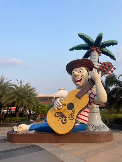 The image shows a large cartoonish statue of a man with exaggerated features, wearing a cowboy hat and a striped shirt. He is holding a yellow guitar and standing next to a stylized palm tree. The statue is located in Kasarvadavali, India. In the background, there are other trees, buildings, and a blue sky. The statue is standing on a platform with a gray and brown patterned surface.