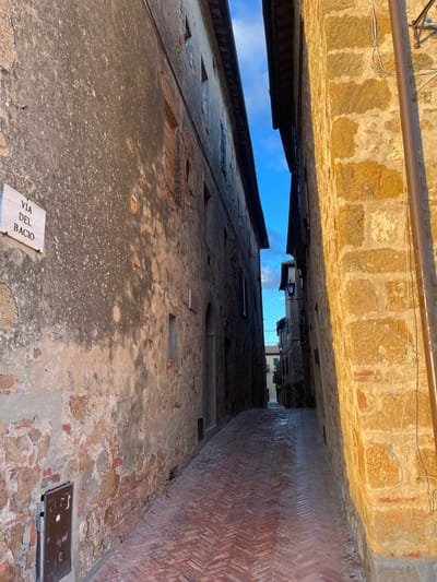 The image captures a perspective looking up a very narrow, historic alleyway in Pienza, Italy. The scene is dominated by the tall, ancient building walls on either side, which rise steeply, leaving only a slender, bright blue strip of clear sky visible at the top center of the frame.

On the left, a weathered building wall is primarily covered in light-colored plaster, though sections of exposed brickwork are visible, indicating its age. Several rectangular cutouts or filled-in window openings punctuate this wall. Towards the bottom of this left wall, a white rectangular sign with black text clearly reads "VIA DEL BACIO," which translates to "Kissing Street" or "Street of the Kiss."

The right side of the alley is defined by another old building wall, featuring a distinctive section of red brick laid in a herringbone pattern, particularly prominent in the lower parts. Higher up, this wall also shows a mix of brick and stone construction. A dark metal drainpipe runs along the upper section of this wall. The "ground" surface of the alley, visible in the lower right, appears to be a worn mixture of plaster and exposed stone, with two dark, square utility covers embedded in it.

The time of day is clearly daytime, with bright sunlight illuminating the upper portions of the buildings, casting a warm, golden glow on the stone and plaster. Conversely, the lower parts of the alley and the ground are in deep shadow, emphasizing the narrowness and height of the passage. No people are visible, and no specific activity is taking place, contributing to a serene and timeless atmosphere. In the distance, at the apparent end of the alley, hints of further buildings and a touch of greenery can be discerned.