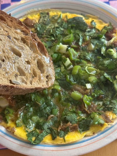 This close-up image captures a meal served on a striped ceramic plate, likely breakfast or brunch. The main subject is an oval-shaped egg dish, resembling an omelet or a similar fried egg preparation, occupying the majority of the plate. It is generously topped with a mixture of finely chopped green herbs, including bright green scallions or spring onions and dark green cilantro or parsley. A dark, glossy savory sauce is also visible, coating the herbs and egg.

Partially overlapping the egg dish in the foreground, and extending from the bottom left, is a thick slice of artisanal bread, possibly sourdough, characterized by its open, airy crumb and visible seeds or grains. The bread appears to be toasted or lightly grilled and has a slight sheen, suggesting it might be buttered or drizzled with oil.

The plate itself has a white base with thin stripes of blue, orange, and white around its rim. The background suggests a wooden surface, indicating an indoor setting such as a home dining table or kitchen counter. No specific time of day or weather is discernible from the image, but the food items suggest a morning or midday meal. There is no visible text in the image. The meal is taking place in Egkomi Lefkosias, Cyprus.
