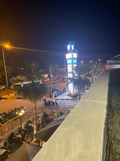 This is a night-time, high-angle shot of a street scene in Yunus Emre, Turkey. A tall, illuminated signpost with multiple screens displays the word "BOYNER" multiple times and abstract graphics. Several palm trees are present. In the foreground, a light-colored concrete wall runs along the bottom right of the frame. Below this wall, on a lower level, a group of people are seated at tables, some under umbrellas. To the left, a street is visible with parked motorcycles and cars. A bus is also seen driving on the street. The sky is dark, indicating it is nighttime, with streetlights and the illuminated sign providing the primary light sources.