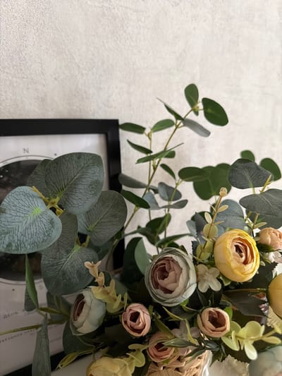 The image shows a close-up view of a floral arrangement in a wicker basket. The arrangement features a variety of artificial flowers in shades of pink, yellow, and light blue, interspersed with green eucalyptus leaves and stems. A black-framed picture is partially visible on the left side of the image, behind the foliage. The background is a textured, off-white wall. The lighting suggests an indoor setting. There are no people visible in the image. The location is specified as Semiluki, Russia, however, there are no visual cues in the image that confirm or indicate this specific location.