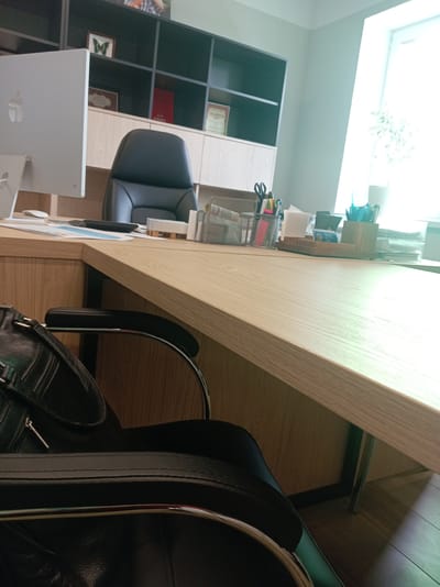 The image shows an office environment. In the foreground, a black leather office chair with chrome accents is partially visible, along with a black leather handbag on the seat. A light-colored wooden desk occupies the center of the frame, with a computer monitor, keyboard, and documents on its surface. Behind the desk, a dark grey shelving unit is mounted on the wall, displaying various items including a framed butterfly specimen, a framed certificate, and what appear to be books or binders. A large window with bright daylight is visible on the right side of the image, illuminating the room. The flooring appears to be wooden planks. The scene is likely located in an office space within a building in the Шчомысліцкі сельскі Савет, Belarus.