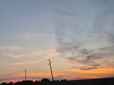 This is a medium shot, eye-level, outdoor photograph taken during twilight. The scene depicts a rural landscape with a silhouette of trees and vegetation along the bottom of the frame. Power lines stretch horizontally across the mid-ground, supported by wooden poles. One pole, located slightly left of center, is more prominent, leaning slightly. In the background, the sky is a gradient of colors, transitioning from a pale blue at the top to warm orange and pink hues near the horizon, indicative of a sunset. Wispy clouds are scattered across the sky, with some appearing as faint streaks from contrails. The overall impression is one of a tranquil evening in a natural setting. The specific location in Churek, Bulgaria is noted.
