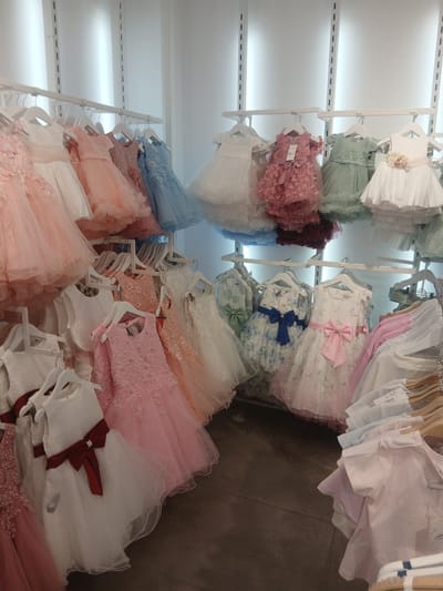 A retail display of children's formal dresses is shown in this image. The dresses are hung on racks and are of various colors, including pink, blue, white, and green. Some dresses are adorned with embellishments such as lace, beads, and bows. The display appears to be in a store, with racks of clothing visible in the background. The lighting suggests an indoor environment. The image does not contain any identifiable people or specific architectural features that would indicate a location in the city of Ávila, Spain.