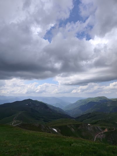 This image captures a wide-angle mountainous landscape in Sedlačka gora, Montenegro, under a predominantly overcast sky. The upper approximately two-fifths of the frame are dominated by dense, grey cloud formations, with several irregular patches of clear blue sky visible through breaks, particularly in the upper right and mid-left, where brighter light penetrates.
The lower portion of the image displays a vast expanse of rolling, verdant mountains and hills. The immediate foreground features lush green grass covering a gentle slope. In the midground, multiple layers of green-clad peaks and valleys recede into the distance. Winding dirt tracks or unpaved roads are visible traversing the hillsides, with one section on the left exhibiting a wooden fence or barrier running alongside it. Several distinct, small patches of snow are present on the green slopes. The distant mountains appear in lighter, desaturated shades of blue-grey, characteristic of atmospheric perspective, blending into the horizon. No people, specific subjects, or interactions are visible. The environment is natural and rugged, indicative of a high-altitude mountain region.