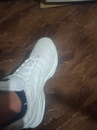 The scene is an indoor setting in Krivodol, Bulgaria. A single, clean white athletic shoe, consistent in design with a Nike Air Max Plus model, is prominently positioned on a dark brown floor with an irregular, light-colored marble or veining pattern. The shoe features a mesh upper with distinct wavy white overlays, white laces, and a thick white sole. A black ankle sock is visible on a human foot, with a portion of the skin of the lower leg/ankle exposed above the sock. The image is captured from a slightly elevated, downward-angled perspective, focusing on the shoe and the immediate floor area. An indistinct light-colored object is partially visible in the upper background.