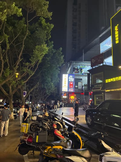 The image depicts a nighttime street scene in Longta, China, characterized by a mix of pedestrian and vehicular activity. Tall trees with dense foliage line the left side of the street, their branches illuminated by streetlights. The street itself is paved and appears wet, suggesting recent rain.
Numerous parked motorcycles and scooters are visible in the foreground and midground, some with storage boxes attached. A black SUV is parked on the right side of the frame, partially obscuring the view of a building with illuminated windows.
Several individuals are present. On the left, a person wearing a striped shirt and light-colored pants stands near parked vehicles. Further down the street, a group of people are gathered, some walking and others standing. A man in a dark shirt and shorts is standing on a raised platform on the right, looking towards the street.
Buildings flank the street, featuring various illuminated signs. One sign, in yellow Chinese characters, reads "影院体验" (Cinema Experience). Another sign, in red and white, displays "茶坊" (Tea House). There are also advertisements and graphics displayed on the building facades. The overall atmosphere is that of a lively urban area at night.