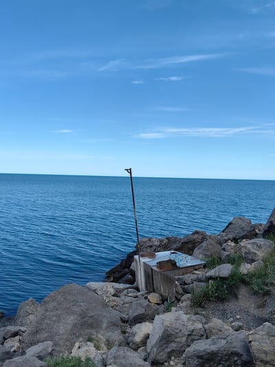 The image captures a serene coastal scene in Chaika, Ukraine. The vast expanse of the Black Sea dominates the frame, its calm, deep blue waters stretching to meet a clear, pale blue sky punctuated by wisps of white clouds. The foreground is a rugged landscape of large, grey rocks, some smooth and rounded, others jagged and rough, forming a natural embankment. Amidst these rocks, a metallic structure, possibly a utility box or a small shelter, stands weathered and grey. On top of this structure, several plastic bottles of water are neatly arranged, their labels still visible. A thin, dark pole with a hook-like attachment rises from behind the metallic box, its purpose unclear in this context. The overall impression is one of natural beauty with a touch of human presence, hinting at a secluded spot by the sea.