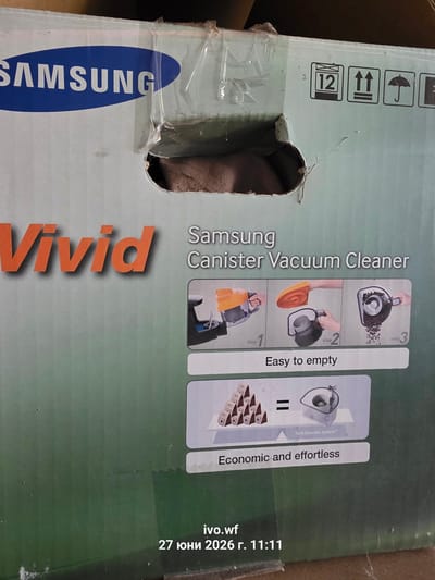 The image shows a cardboard box for a Samsung Vivid Canister Vacuum Cleaner. The box has the Samsung logo in blue and white, and the word "Vivid" in large orange letters. There are also diagrams illustrating how to empty the vacuum cleaner, labeled "Easy to empty" and "Economic and effortless." Shipping symbols are visible on the top right of the box. The box appears to be damaged in the upper left corner. The bottom of the image contains text that reads "ivo.wf 27 юни 2026 г. 11:11". The exact location within the city of Chal, Bulgaria is not provided.