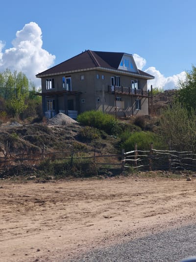 A wide-angle, daylight shot captures a two-story house situated on a hill under a bright blue sky dotted with large, fluffy white cumulus clouds.

In the foreground, a wide, sandy dirt road with visible tire tracks and scattered small rocks stretches across the bottom of the frame, transitioning into a gravel surface on the far right. A rustic wooden fence, constructed from irregularly shaped branches and logs, runs horizontally across the midground, delineating the road from a rising, uneven slope.

The house, the central subject, features a dark brown hip roof with light-colored eaves and neutral grey or beige stucco-like walls. It has multiple windows, some with white frames, and several balconies with decorative dark metal railings. A distinct dormer window with blue-tinted panes is visible on the upper roof section. On the lower level, a pile of light-colored sand sits next to what appears to be a covered porch area, suggesting ongoing construction or landscaping.

The terrain around the house is hilly and somewhat arid, covered with dry grass, sparse green bushes, and some taller, vibrant green trees, particularly on the left and right sides of the slope. The scene is brightly illuminated by the sun, indicating a clear day. No people or visible text are present in the image. The overall setting appears to be a rural or semi-rural landscape, consistent with the location context of Kiçi-Jargılçak, Kyrgyzstan.
