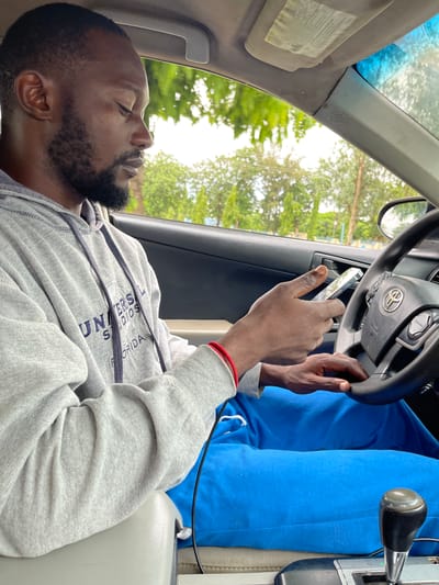 A dark-skinned man with a beard is sitting in the driver's seat of a car, looking at his phone. He is wearing a gray hooded sweatshirt with "UNIVERSAL STUDIOS FLORIDA" printed on it in navy blue lettering, and bright blue sweatpants. A red bracelet is visible on his left wrist. The car interior is visible, including the steering wheel with a Toyota logo, a gear shift, and part of the dashboard. Outside the car, trees and foliage can be seen, suggesting an outdoor environment. The scene is likely within the Immigration Quarters, Nigeria, given the context provided, indicating a likely urban or semi-urban setting in Nigeria.
