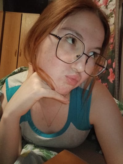 The scene is an indoor portrait of an adult female, identified as occurring in the city of Старый Шагирт, Russia. The subject is depicted from the upper chest, with reddish-brown hair and a light complexion, exhibiting minor facial blemishes. She wears large, black-framed, hexagonal eyeglasses and a light grey tank top with turquoise blue shoulder and neckline trim. A delicate gold-colored chain with a small pendant is visible around her neck. Her left hand supports her chin, and her lips are pursed. Her gaze is directed upward and to the right of the frame. The background to the left consists of a light brown wooden two-door cabinet or wardrobe. To the right, a floral patterned wallpaper or curtain, featuring pink/red flowers, is visible. The lower foreground is composed of a blurred, patterned textile.
