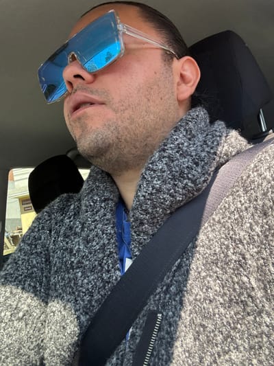 A male individual is observed seated in the front passenger compartment of a vehicle in San Juan Cuautlancingo, Mexico. He wears large, clear-framed sunglasses with reflective blue lenses, which show reflections of power lines and tree foliage. The subject has short facial hair, and his dark hair appears pulled back. He is dressed in a grey, textured, collared sweater with a visible zipper closure, layered over a blue garment. A black seatbelt is fastened across his chest and left shoulder. The vehicle's interior features a black headrest. Outside the vehicle, visible through a side window, is a light-colored building with a distinct yellow section.