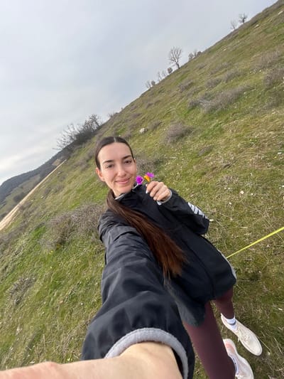 A single adult female is depicted outdoors in Ostar Kamak, Bulgaria. The subject is positioned facing the camera, with a faint smile, and her left arm is extended towards the lens. Her long, dark brown hair is pulled back into a ponytail. She is dressed in a black zippered athletic jacket with light grey accents on the sleeves, dark reddish-brown leggings, and white athletic shoes with pinkish details. In her left hand, she holds two small flowers, one predominantly purple and the other yellow-orange. The background consists of a sloping hillside covered in green and dry grassy patches, with sparse, bare deciduous trees and shrubs. A dirt track or road is visible along the base of the hill on the far left. The sky is uniformly overcast. A yellow string or rope is visible on the ground, stretching from the lower right foreground towards the center-right midground.