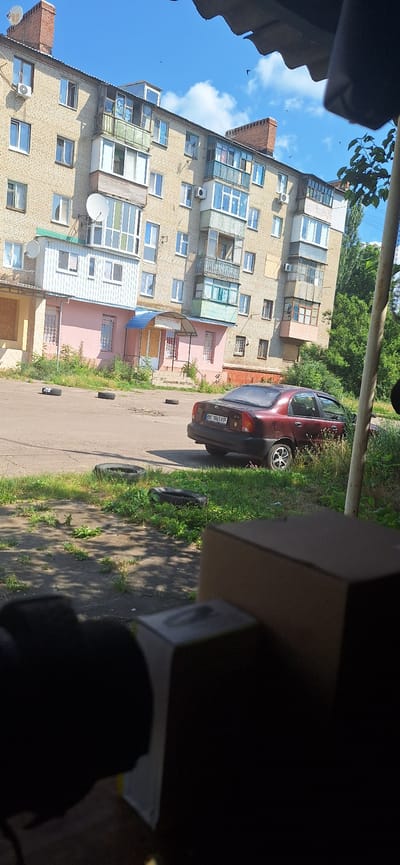 The image depicts an exterior view of a multi-story apartment building, likely in Sloviansk, Ukraine. The building is constructed of light-colored brick, with multiple windows, balconies, and satellite dishes visible. The ground floor features a pink-colored section with a covered entrance labeled "ГАСТРОНОМ" (Gastronome). In front of the building, on a paved surface, there are several discarded car tires. A maroon sedan is parked on the right side of the image, facing away from the camera. The area is overgrown with grass and vegetation. The sky is blue with scattered clouds, and several birds are visible flying. In the foreground, a blurry, out-of-focus view of what appears to be the corner of a cardboard box and possibly a camera lens is present.