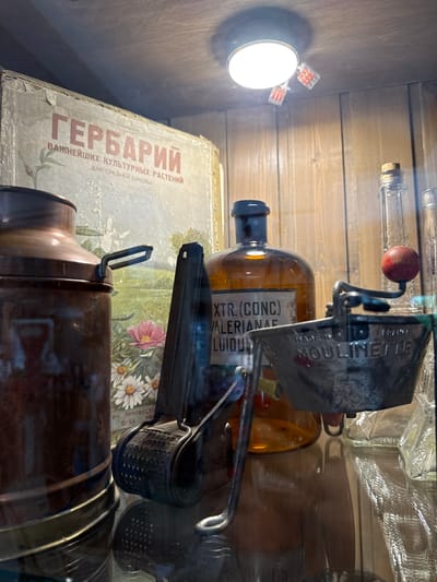 The image depicts a close-up view of items displayed on a shelf in what appears to be a museum or an antique shop. The backdrop consists of wooden paneling. In the foreground, a collection of objects is visible. To the left, a large, tarnished copper container with a handle is partially seen. Adjacent to it, a vintage manual food grinder, likely a Mouli or similar brand, with a red wooden knob is positioned. Behind these items, an amber-colored glass bottle with a dark cap is prominently featured. This bottle has a label that reads "EXTR. (CONC.) VALERIANAE FLUIDUM" and also has some text in Russian which includes "ВАЖНЕЙШИХ КУЛЬТУРНЫХ РАСТЕНИЙ" and "ДЛЯ СРЕДНЕЙ ШКОЛЫ" on a faded book or poster in the background, suggesting a connection to botanical studies or educational materials. The book or poster itself shows a painted landscape with flowers, including daisies and a pink rose. To the right of the amber bottle, several clear glass bottles of various sizes are stacked. A small circular light fixture is mounted on the ceiling, with two red wire connectors attached to it. There are no people visible in the frame. The overall impression is of a curated display of historical and scientific artifacts. The provided location of Semogorskiy, Russia, is not discernible from the visual information presented in the image.