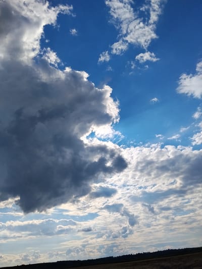 The image displays a dramatic sky over a rural landscape, likely in Ustyvytsya, Ukraine. Dominating the left side of the frame is a large, dark, ominous cloud, heavily shadowed and textured. To the right, the sky transitions to a vibrant, clear blue, dotted with smaller, scattered white clouds, some of which are wispy and illuminated by sunlight. Beams of light appear to emanate from behind the dark cloud, creating a stark contrast and a sense of impending weather change. The horizon line, visible at the bottom of the image, shows a dark silhouette of a gently rolling terrain, possibly fields and distant trees or a small cluster of buildings. The overall impression is one of a dynamic and powerful natural display. No people, specific objects, or actions are discernible.