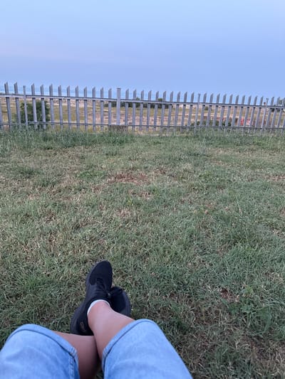 A low-angle, wide shot captures a person sitting on a grassy field next to a fence. The person's legs are crossed, wearing blue denim jeans and black sneakers with white socks. The field is covered in green and brown grass, with some patches of dry earth visible. A metal fence with vertical bars and horizontal supports runs along the left side of the frame. Beyond the fence, a hazy landscape can be seen, with a pale blue sky overhead, suggesting it is either early morning or late afternoon. The overall mood is relaxed and peaceful. No visible text is present in the image. The location context is Shkorpilovtsi, Bulgaria.