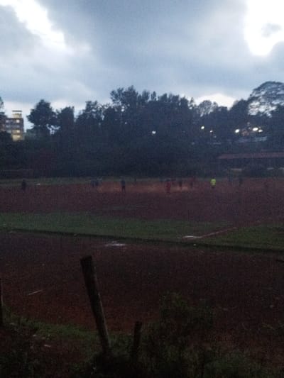 The image shows a view of a dirt field from a slightly elevated position. The field is located in Othaya, Kenya, at coordinates -0.44 latitude, 36.959 longitude. The field appears to be a sports field as there are a group of people playing soccer, with 11 figures visible in the distance. There are trees in the background and the sky appears to be overcast. The scene is shot in the late afternoon or early evening as the sun is casting a long shadow. The image is slightly blurred, as if taken with a phone camera.