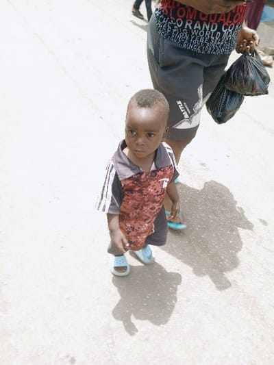 The scene depicts a young, dark-skinned male child and an accompanying adult on an unpaved, light-colored ground surface in Kadoma, Zimbabwe. The child, positioned slightly left of center, wears a short-sleeved collared shirt with a red-brown camouflage pattern on the front and dark-and-white stripes on the shoulders, paired with light blue open-toed sandals. His short, dark hair is visible, and he gazes slightly to his right. Behind and to the right of the child, the lower torso and legs of an adult are visible. This adult wears dark grey shorts with white graphics, including text like "BATTLE," and a red-and-black patterned shirt with various white text elements such as "WORLD" and "BRAND." The adult holds two black plastic bags in their left hand and wears light blue open-toed sandals on their visible foot. Several blurry figures are present in the distant background. The environment is brightly illuminated by daylight, casting strong, distinct shadows of the subjects onto the ground.