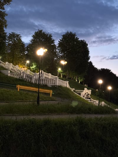 This media file captures a park scene in Vladikavkaz, Russia, during twilight. The sky is a deep blue, with hints of lighter hues near the horizon, suggesting the transition from day to night. Dark clouds are present, adding to the dramatic atmosphere.

The primary subject is a long, ornate white stone staircase that winds its way up a grassy slope. Several lampposts with bright, warm lights illuminate the path and the surrounding area, casting a glow on the white railings and steps. Trees with dark foliage flank the staircase, their branches silhouetted against the darkening sky.

There are a few people visible on the staircase. In the upper portion of the image, a group of individuals appears to be seated or standing near the top of the stairs, seemingly enjoying the evening. Further down, a single person is captured walking or sitting on the stairs. The overall mood is peaceful and serene, with the lighting creating an inviting ambiance in the park.

A yellow bench is placed alongside the lower part of the staircase, facing away from the camera. The grass on either side of the paved path and staircase is lush and green, with some areas appearing darker due to the diminishing light. The scene suggests a public park or garden area, popular for evening strolls and relaxation. No visible text is present in the image.