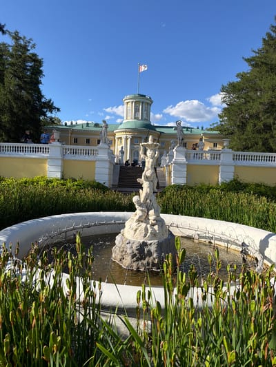 The image captures a scene at Arkhangel'skoe, Russia, on a bright, sunny day. The focus is on a section of the grand estate, featuring a building with pale yellow walls and a green roof. Architectural details include white balustrades, columns, and arched windows. A notable element is a domed structure topped with a small tower, from which a flag flies.
In the foreground, a white fountain with a weathered stone sculpture at its center is partially visible. The water from the fountain creates a delicate spray, and the basin is surrounded by tall, green reeds. The image is oriented vertically, with the sky taking up a significant portion of the upper left quadrant, filled with scattered white clouds.
Several people are present, though their details are somewhat obscured. Some are seen standing on a balcony and on the stairs leading up to the building, observing the surroundings. The overall atmosphere appears peaceful and relaxed, indicative of a leisurely visit to a historical site. The bright sunlight suggests it is daytime, likely late morning or afternoon. The lush greenery and clear sky contribute to a pleasant, summery ambiance. No visible text is present in the image.