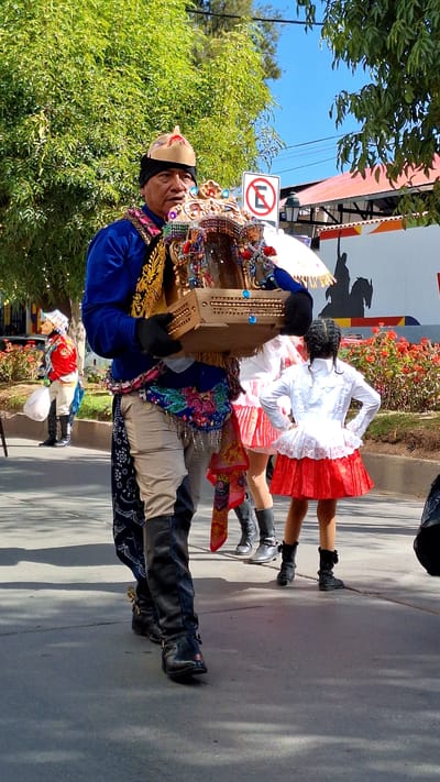 In the city of Wanchaq, Peru, a man in traditional clothing, featuring a blue jacket, tan pants, and elaborate decorative sash, walks forward carrying a gilded, ornate miniature structure. He wears a golden headdress with red accents and black gloves. Behind him, two young girls are visible. One, in the foreground, wears a white blouse and a red pleated skirt, with her back to the camera. Another, further back and to the left, is dressed in a red and white traditional outfit. The background shows a street with trees, flowering bushes, and a building with a red roof. A "No Entry" sign is also visible. The lighting suggests it is daytime.