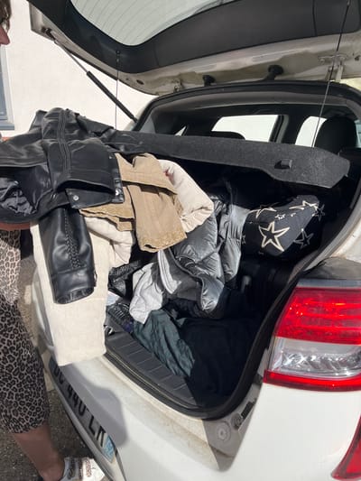 The image shows an open car trunk filled with various items. Predominantly, there are several pieces of clothing including a black leather jacket, beige jacket, a light-colored corduroy garment, a grey quilted jacket, and a black bag with white stars.

The trunk belongs to a white car. The tail light of the car is visible on the right. The license plate reads "DC-440-LK".

The location is Montélimar, France, based on the license plate number. A person's legs, wearing leopard print pants and white sneakers are visible beside the trunk.