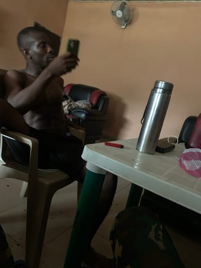 An indoor scene in Kado, Nigeria, features an adult male with a fade haircut, shirtless and wearing dark shorts, seated in a light-colored plastic chair on the left. He holds a green-cased smartphone in his right hand, looking at the screen. His left arm rests on the chair's armrest. A white plastic folding table with green legs occupies the foreground. On the table, a silver metallic thermos flask with a black strap stands upright, alongside a red lighter, a small dark rectangular object, a white translucent plastic bag with pink trim, and a white circular object. The lower foreground shows the legs of another individual, clad in green compression socks and camouflage-patterned trousers, seated opposite the first man at the table. The background wall is painted a light orange, featuring a white wall-mounted oscillating fan in the upper right. A black and red upholstered armchair is partially visible in the left background. The floor is tiled.