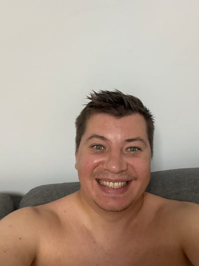 A man is captured in a selfie with a light grey wall and a grey couch visible. He is smiling broadly, revealing his teeth, and his eyes are open, looking towards the camera. His hair is dark and styled upwards. He is shirtless, and his skin appears slightly shiny. The lighting suggests it could be daytime. The background is neutral, with no specific objects or prominent details that indicate a particular location beyond the general setting of an indoor space. The man's expression conveys happiness or amusement. There is no visible text in the image.