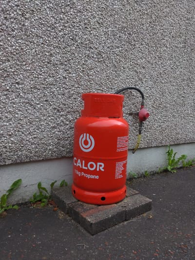An outdoor, eye-level, medium shot captures a red Calor 11kg propane gas cylinder standing upright on two grey concrete blocks next to a textured, pebble-dash wall. A black gas hose extends from the top of the cylinder, connecting to a red gas regulator mounted on the wall with a bracket. The hose is partially visible, with a yellow band near the regulator. Small green weeds grow from cracks in the asphalt ground around the base of the cylinder and the wall. The wall appears to be part of a building. The scene is devoid of any people or animals. The lighting suggests daylight, and the environment is consistent with a residential or commercial outdoor space. The specific location within Magherafelt, United Kingdom, cannot be determined from the visual information alone.