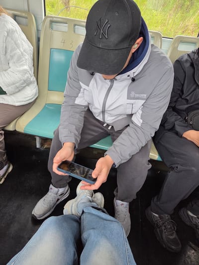The image, taken from an overhead perspective, captures a view inside what appears to be a public transport vehicle, likely a bus or train, with several passengers seated. The primary subject is a person in the foreground, wearing a grey jacket with a dark blue hood, black athletic pants, and a black baseball cap with a white "NY" logo. They are holding a blue smartphone with both hands, looking down at its screen. A light green in-ear device is visible in their right ear. On the jacket sleeve, the brand "FAVORITE" and "ORIGINATED IN 1993" are visible.

To their left, another passenger is partially visible, wearing a white quilted jacket over light-colored pants. Below this person, a pair of blue denim jeans and light-colored sneakers can be seen, possibly belonging to the same individual or a different one. The floor of the vehicle is dark and appears to be a mat.

In the upper right, another passenger is seated, clad in dark, possibly black, clothing and dark sneakers. The seats are light beige with blue-green cushions. On the back of the seat in front of the main subject, the number "43" is written in marker. Outside the window, vibrant green foliage and natural light suggest it is daytime, though the weather appears overcast or diffused. The overall scene depicts individuals traveling, with one focused on their phone. Emotions are not discernible due to obscured faces.
