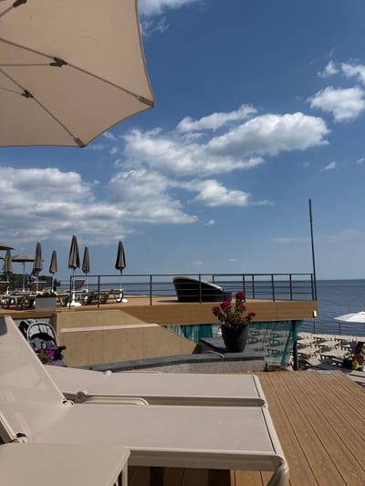 This image depicts a multi-tiered outdoor leisure area situated on the shore of the Kyiv Sea (Kyiv Reservoir) in Hlibivka, Ukraine, under a clear blue sky with scattered white cumulus clouds.

In the foreground, a portion of an open light-beige parasol is visible in the upper left. Two light-beige lounge chairs, positioned on a light-brown wooden deck, occupy the lower half of the frame, facing away from the viewer.

The midground features a terraced layout of light-tan stone or concrete structures and light-brown wooden decking. A black plant pot with vibrant pink/red flowers sits on a ledge to the right. A dark-colored baby stroller is partially visible on a lower deck level to the left. Further back on a deck, several dark-brown folded beach umbrellas and numerous empty lounge chairs are arranged. An elevated wooden deck, bordered by a dark metal railing, features a single dark, curved lounge chair facing the water. A tall, dark pole stands vertically near the right edge of this deck.

The background is dominated by the calm expanse of the Kyiv Sea. Below the elevated decks, a lower beach area is visible, populated by numerous empty light-colored lounge chairs and at least one open white beach umbrella. One individual is seated on a lounge chair on the lower beach near the white umbrella. Another person is partially discernible further left on a deck among the lounge chairs. No specific actions or interactions beyond leisure are clearly depicted.