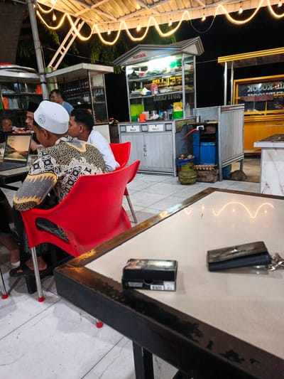 The image captures an outdoor nighttime scene at a food stall in Cotpuuk, Indonesia, illuminated by string lights. In the foreground, a light-colored table is visible with a black wallet and a small black box of cigarettes on it. A portion of a dark-colored table frame is also in view.
The mid-ground features several individuals seated at red chairs around tables. One person wears a white skullcap and a batik-patterned shirt. Another person, to their right, wears a white shirt and also has a skullcap. A laptop is open on a table in front of them. Behind the seated individuals, a food stall with various items displayed on shelves and a counter is visible, with signage indicating "MARTABAK". There's also a propane gas tank near the stall.
The background reveals another food stall with yellow accents and signage that reads "KULINER". The overall environment appears to be a casual outdoor dining area, likely a street food or night market setting. The lighting suggests it is after dark.
